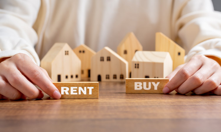 A man is holding two wooden blocks that have the words “RENT” and “BUY” painted on them. Behind his hands is a collection of small wooden house models.