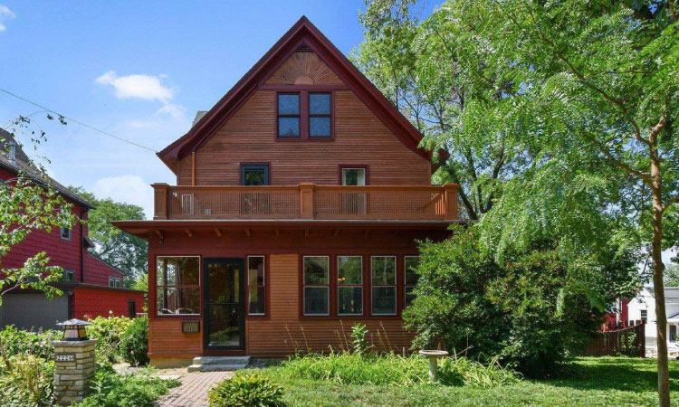 A beautiful two-story, natural wood siding home in the Schenk-Atwood neighborhood of Madison, Wisconsin. The second level features a large enclosed deck, and there’s lovely wood detailing at the top of the gable. The front yard is lush and green.