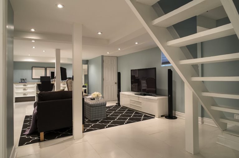 A newly renovated basement, modeled to be an additional living room and home office. The space features an area rug, sofa, and entertainment system in the middle of the room and a desk with an office chair off to the side.