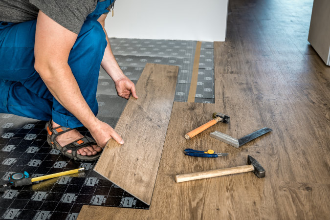 Close up of a person in sandals installing vinyl plank flooring in the basement. 