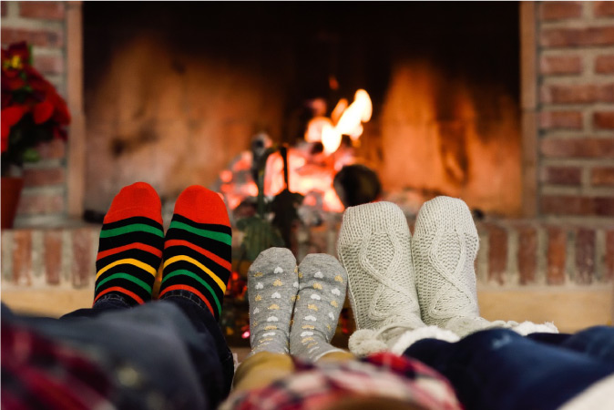 Three pairs of sock-clad feet are being warmed by a fire in a brick hearth.