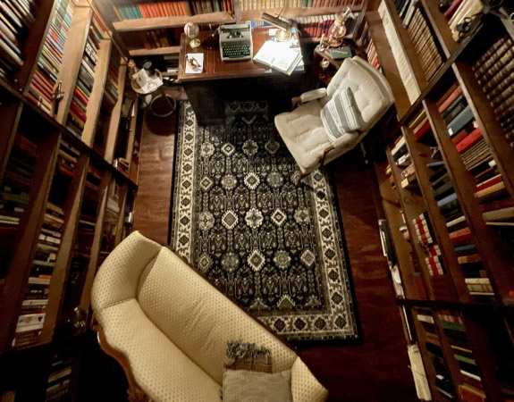 View from above of a home library set up in a basement. The walls are lined with built-in wood shelves that are filled with hundreds of books. There’s a cozy area rug on the floor, as well as a desk, a sofa, and an upholstered chair.