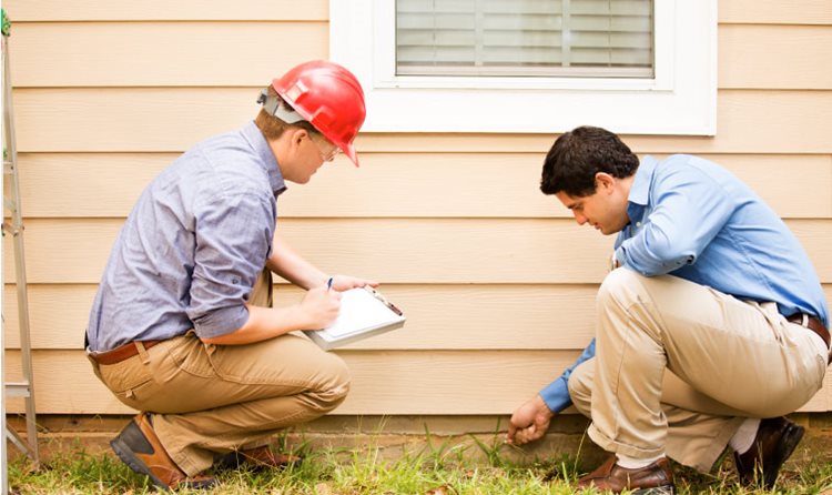 A home inspector and his assistant examine the foundation of a residential home. The inspector is pointing out a crack in the concrete beneath the siding.