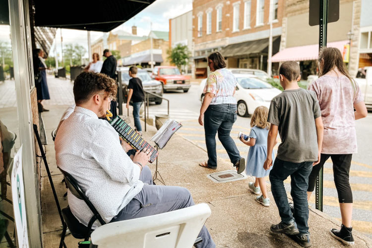 On a sunny afternoon in downtown Batesville, Arkansas, a young man wearing a smart button-up shirt and slacks plays a melodica on the sidewalk. A passing family enjoys the music as they stroll by. Bustling streets and sidewalks create a lively atmosphere as locals explore and engage with their vibrant town.