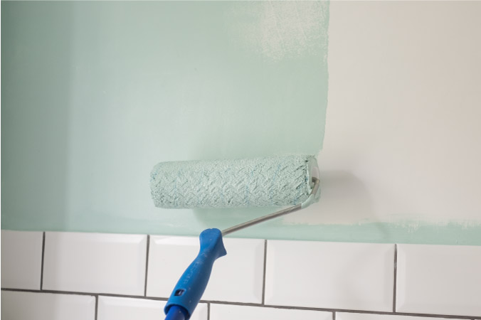 Close-up of a paint roller being used to apply light blue paint to a bathroom wall during a bathroom remodel project