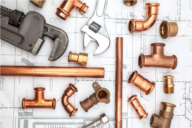 An array of pipes, wrenches, and pipe fittings are  resting atop plumbing plans