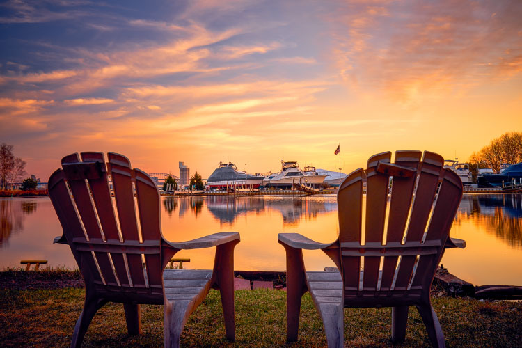 Two Adirondack chairs are set up on the bay side of Park Point in Duluth, Minnesota. The sun is setting across the water, and there are several boats dry-docked across the way.
