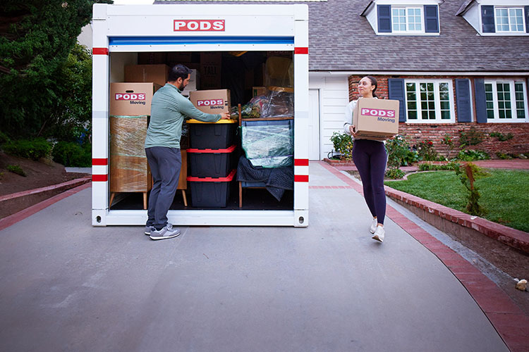 A couple load their PODS container in their driveway. A man loads a cardboard box on a stack of totes and a woman carries a cardboard box to the front of the container