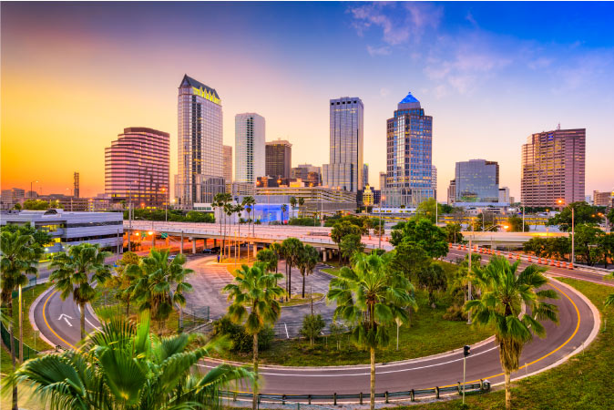 View of the Tampa city skyline at sunset from behind a freeway ramp and several tropical palm trees.