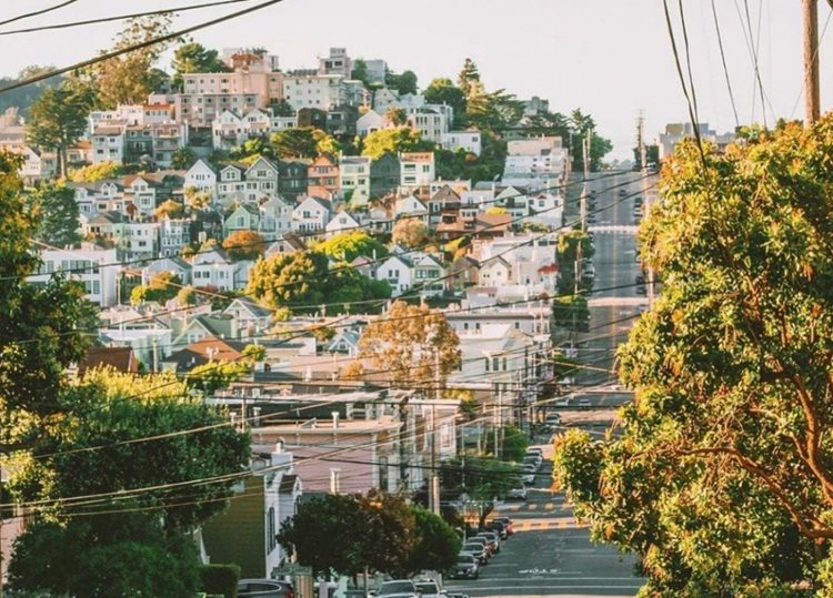 The view up a steep road in the Castro neighborhood of San Francisco. The hill to the left of the road is filled with typical San Francisco homes.