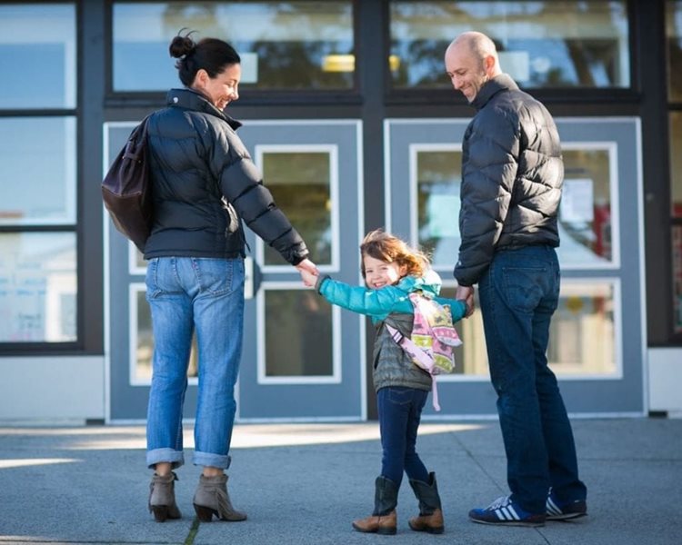 A young girl wearing a school backpack is looking back as she walks into her San Francisco school, holding hands with both of her parents. 