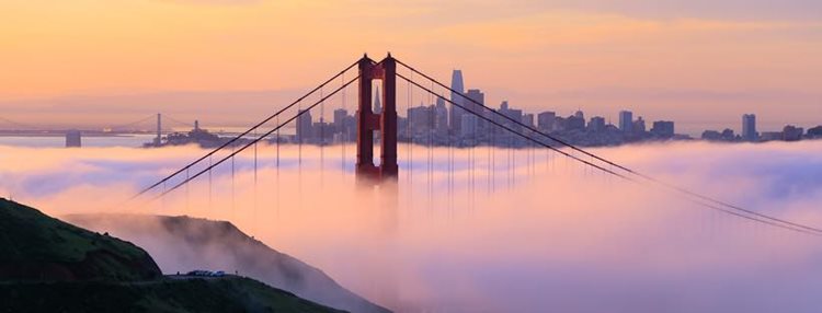 Distant view of Golden Gate Bridge engulfed in fog just after sunset. Downtown San Francisco peaks up above the fog in the distance.