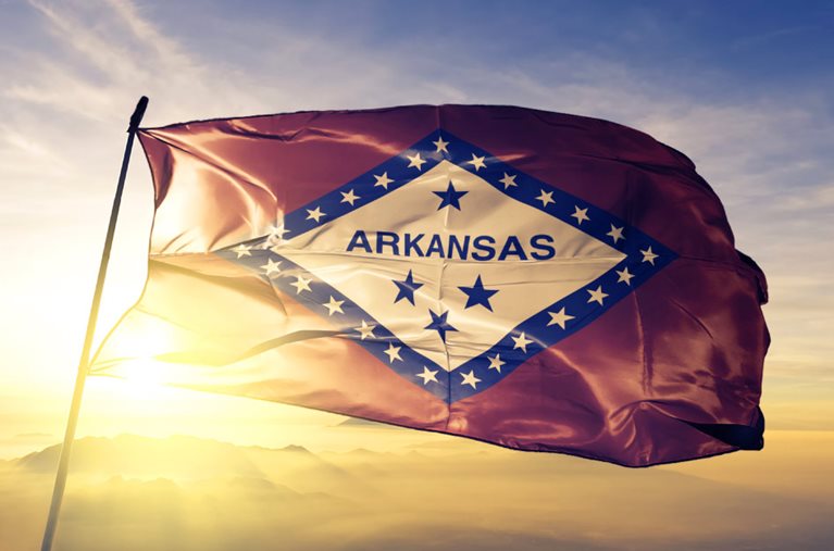 The Arkansas state flag is fluttering gracefully in the wind atop a flagpole. The flag features a red field with a large white diamond in the center. In the diamond, the word “Arkansas” is written in all caps, with many small white stars and four large blue stars. Behind the flag, there’s a serene mountain range with the sun setting behind it.