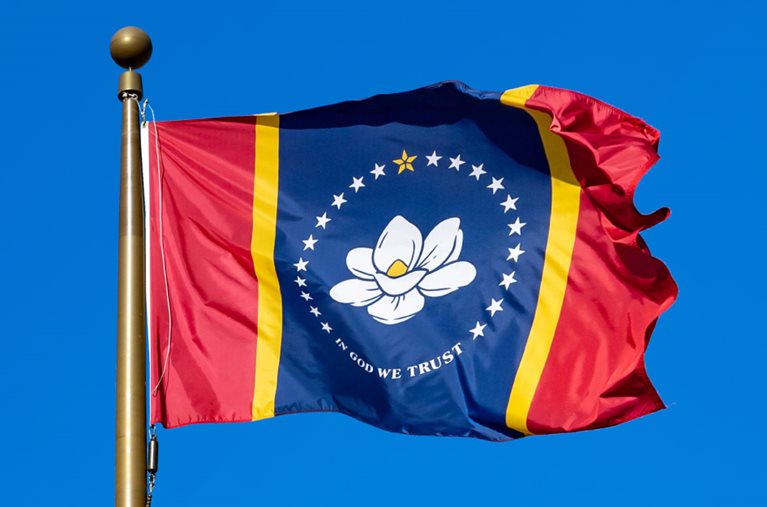 The state flag of Mississippi is blowing in the wind at the top of a flagpole in front of a clear blue sky.