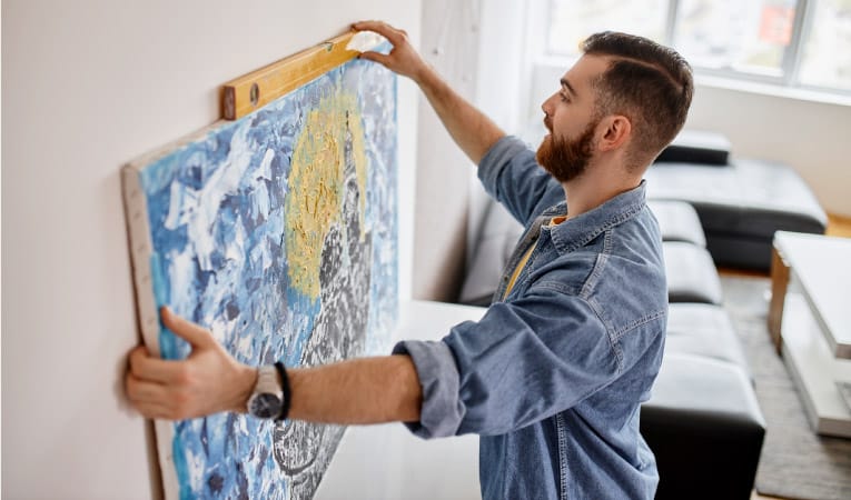 A man is using a level to determine the correct placement of a large painting that he’s hanging in his apartment.