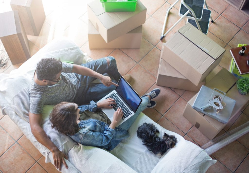 A husband, wife, and terrier sit on a couch, scheduling their moving services on a laptop