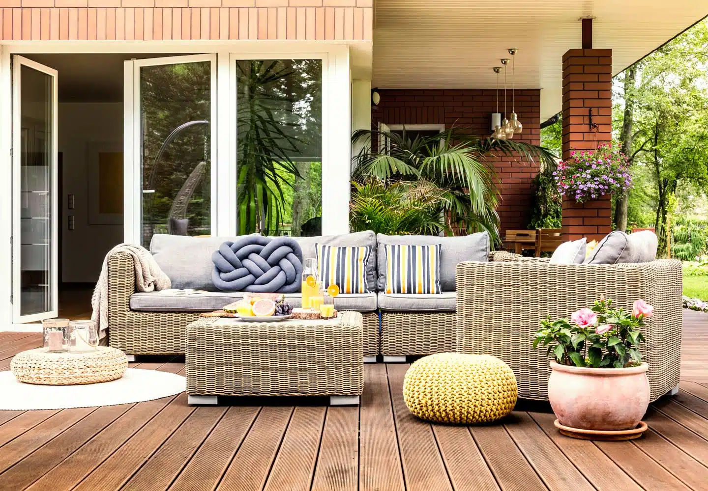wicker furniture sits on the back patio of a home. There is a chair, couch and table. The couch and chair have grey, blue and yellow pillows on them.