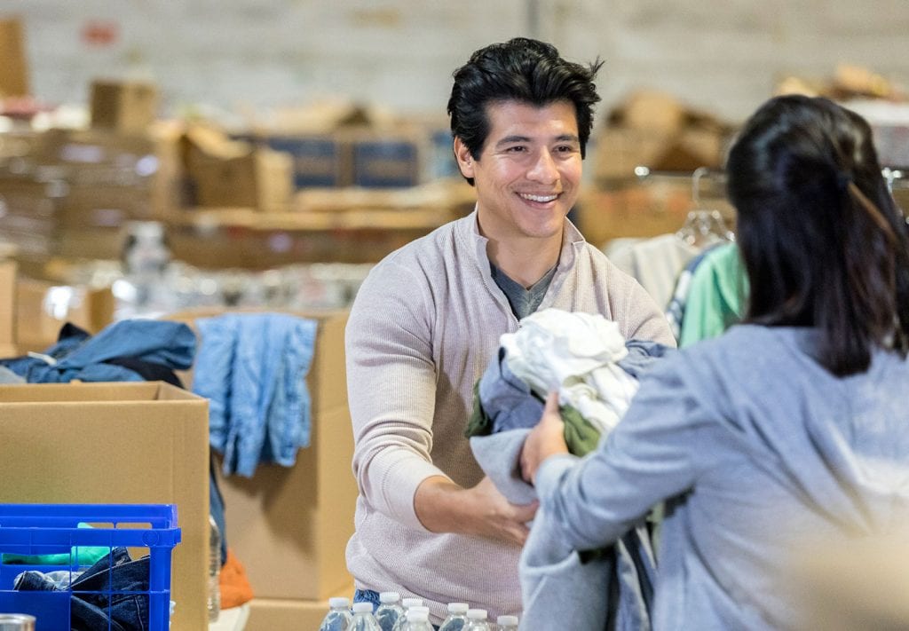 A young man  accepts a dopnation of clothes from someone in a clothing distribution center. Behind him are rows of cardboard boxes. 