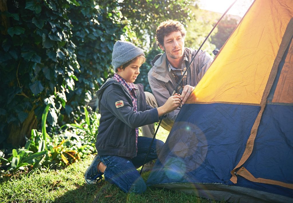 A father and son sit in the forest and set up their yellow tent. 