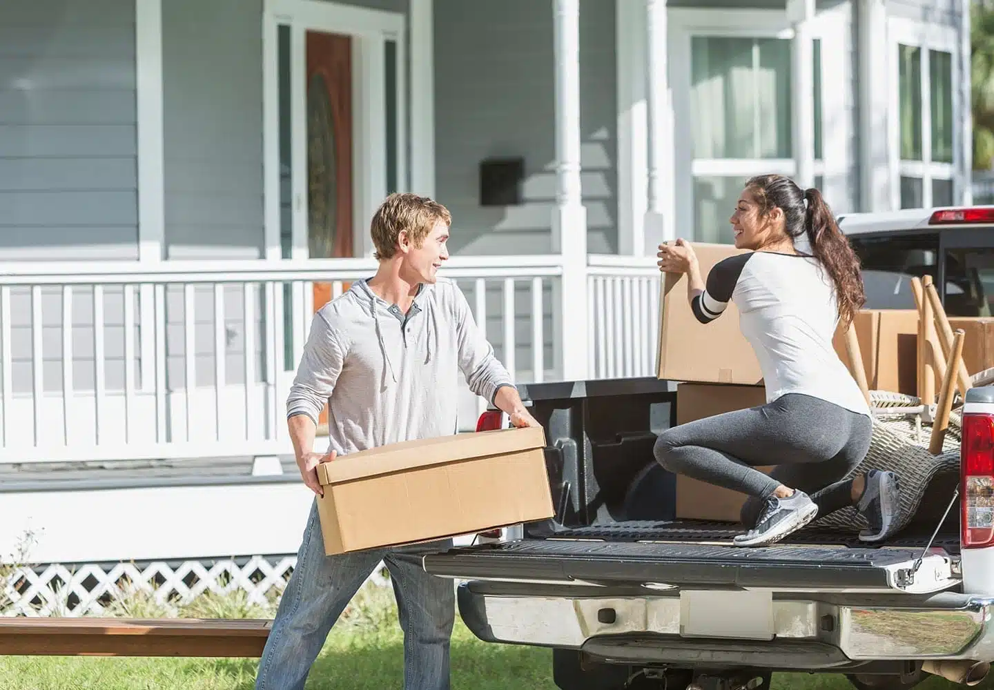 A young couple taking cardboard boxes out of the bed of a pickup truck parked in front of their new home