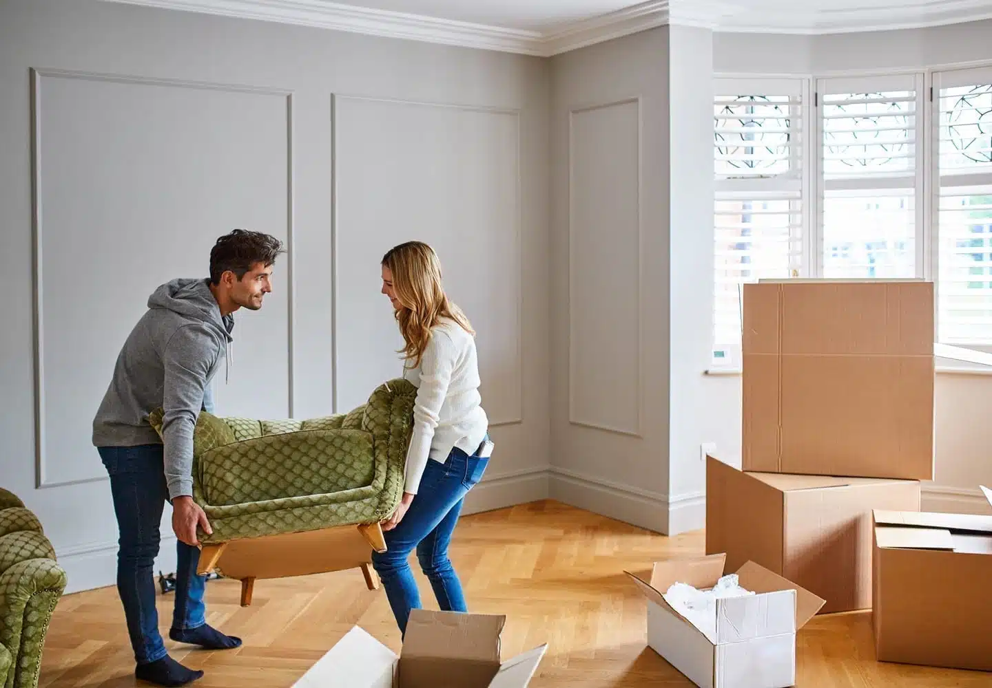 A couple is moving in together. They’re carrying a chair into the living room that’s filled with moving boxes and a matching sofa.