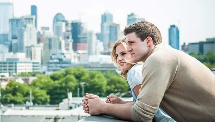 A couple looks out over the city skyline from their balcony. Tall buildings are in the background.