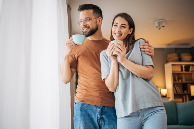 A happy couple standing together by the window after moving in together, looking out at the day as they enjoy their morning coffee.