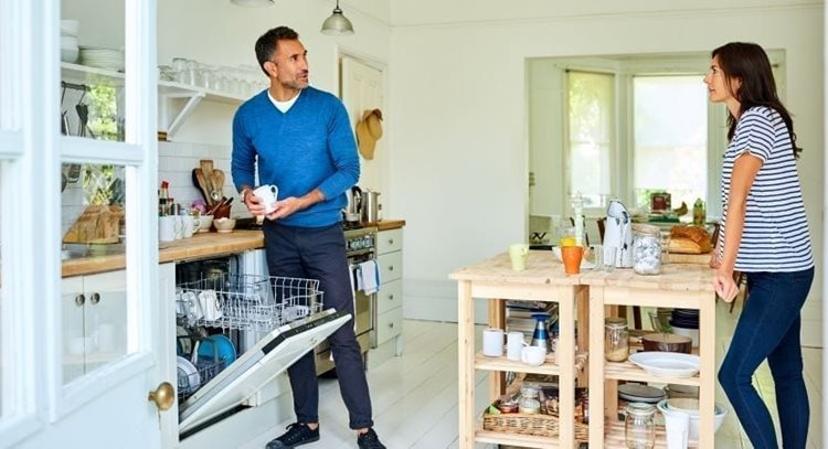 A couple is discussing serious issues in their kitchen. The man is unloading the dishwasher while the woman is leaning against the island.