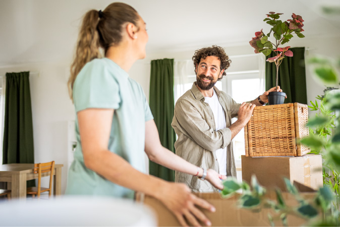 A couple is unpacking boxes while moving in together. The man has brought a plant from his previous apartment to add to the decor of their shared home.