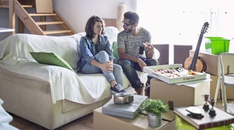 A couple is taking a break from moving in together and eating pizza. They’re sitting on the couch and surrounded by moving boxes and other items that have just been unpacked.