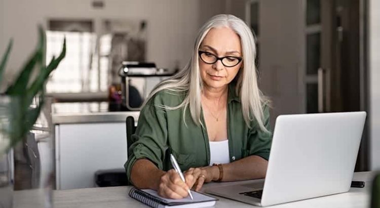 A woman is sitting at the kitchen table with her laptop in front of her doing research. She’s writing in a notepad