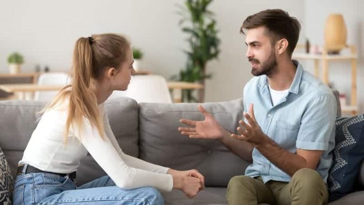 A couple is sitting together on a couch having a deep conversation. They’re both turned toward each other, and the man is raising his hands while he speaks.