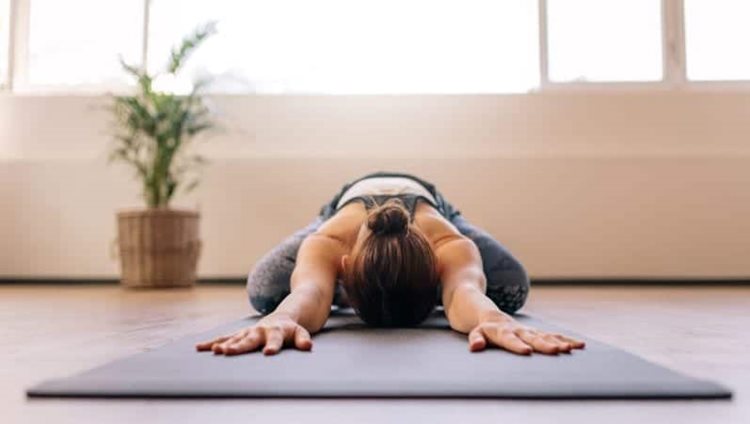 A woman is having some “me time” by doing the child’s pose on a yoga mat. There is a small plant behind her in the background.