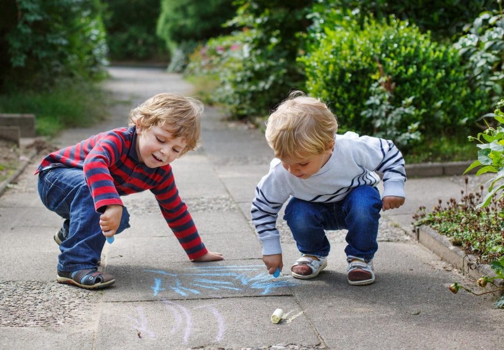 Two young boys play with chalk on the sidewalk. They are smiling. 