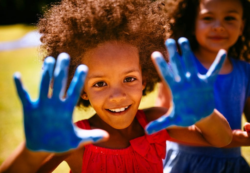 A young girl smiles and shows off her hands that are painted blue. Another young girl smiles behind her. 