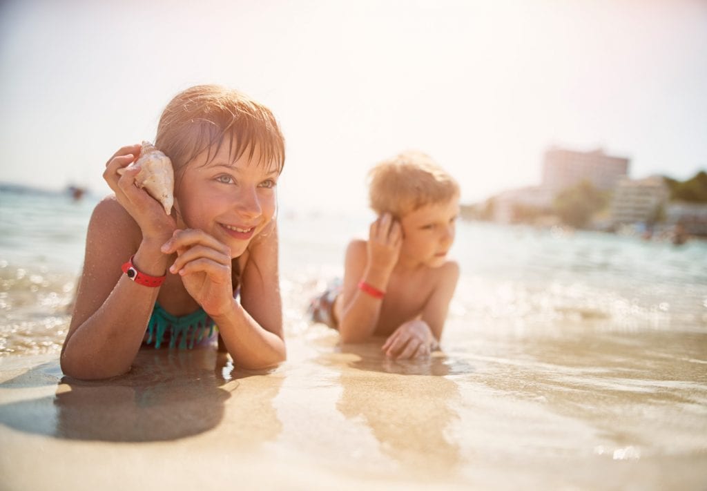 Two kids (a boy and girl) play on a beach, They are holding up seashells to their ear to hear the ocean.