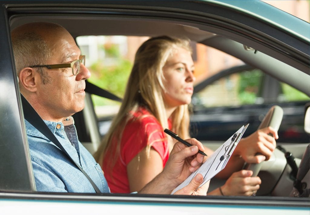 A young, blonde woman sits in the driver's seat of her car. There is a middle-aged man in the  passenger seat checking items off a list as she takes her driving test. 