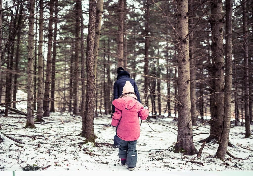 A child in a pink sow jacket and blue snow pants walks behind her mom in a bare forest. Her mom is wearing a navy snow jacket and a black hat. 