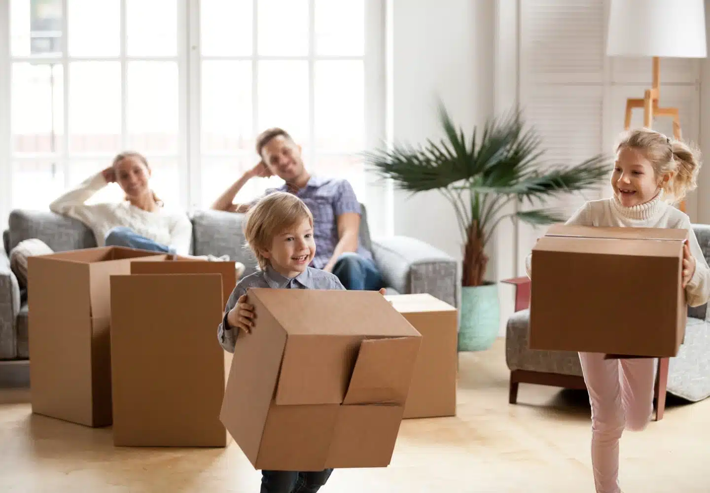 Young parents look on as their kids run in the opposite direction white holding cardboard boxes.