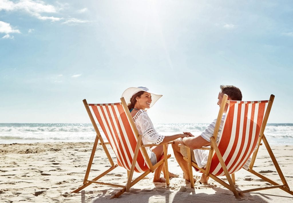 A couple sits in red and white chairs on a sandy beach. They are holding hands, laughing and smiling at each other. 