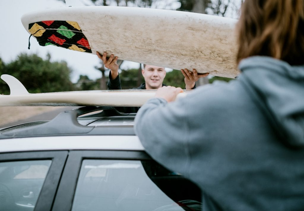 A young man looks at his girlfriend while taking a surfboard off the roof of their shared car. 
