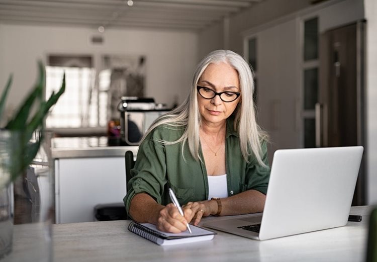 A mature woman with silver hair and glasses sits at a table and takes notes in a notepad. 