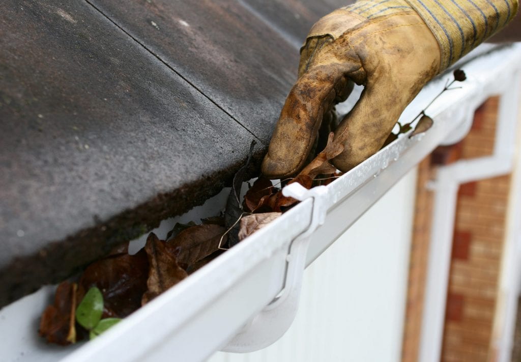 A close up of a dirty glove reaching into a gutter to pull out leaves. The white gutter sits below a black tiled roof. 