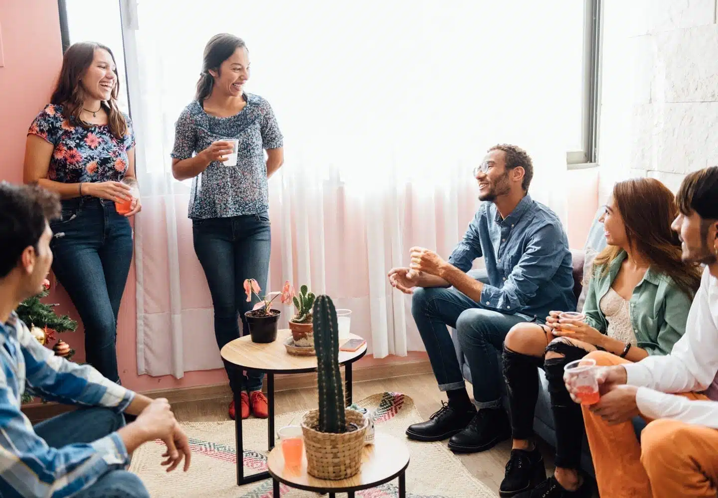 A group of friends sit around at a housewarming party. There are two tables with different plants on them.
