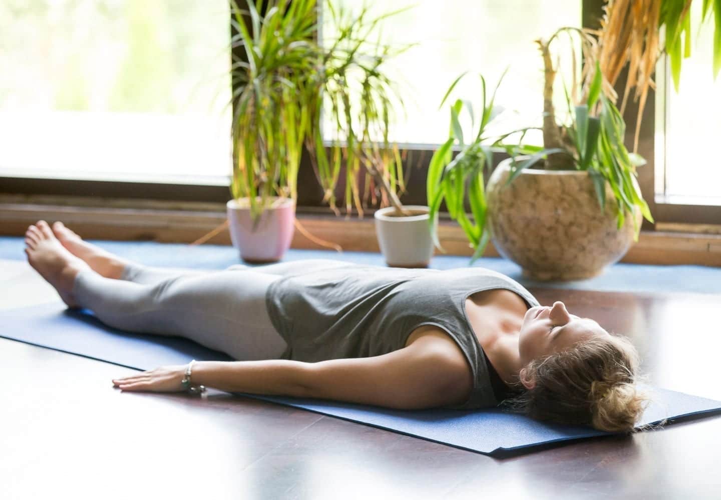 A woman in grey yoga pants and a grey top lays flat on her back practicing the Shavasana pose. 