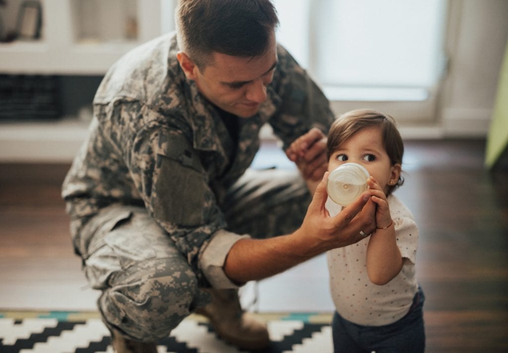 A military man feeds his young daughter a bottle. She looks to the right and her ears are pierced. 