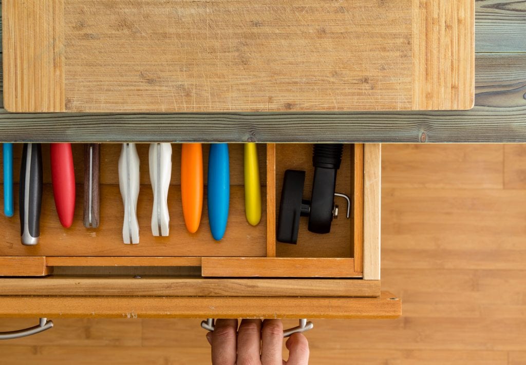 A hand is partially opening a kitchen drawer showing divided storage for knives and a can opener.