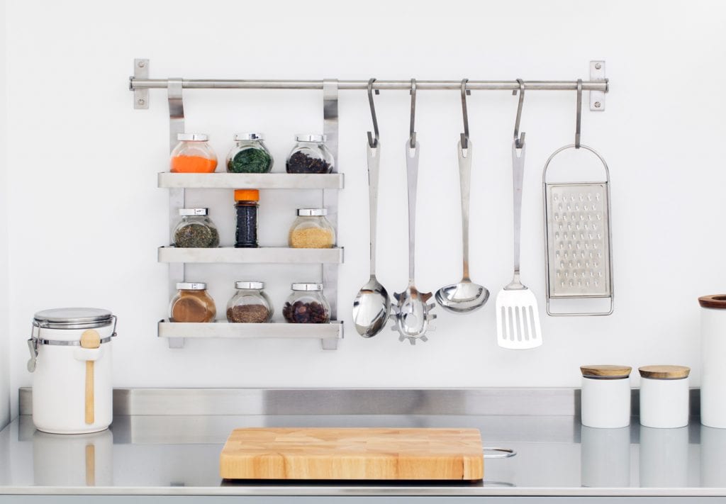 An organized kitchen counter with a few closed canisters and a small butcher block cutting board. A spice rack and cooking utensils are hanging on a rod above the counter.