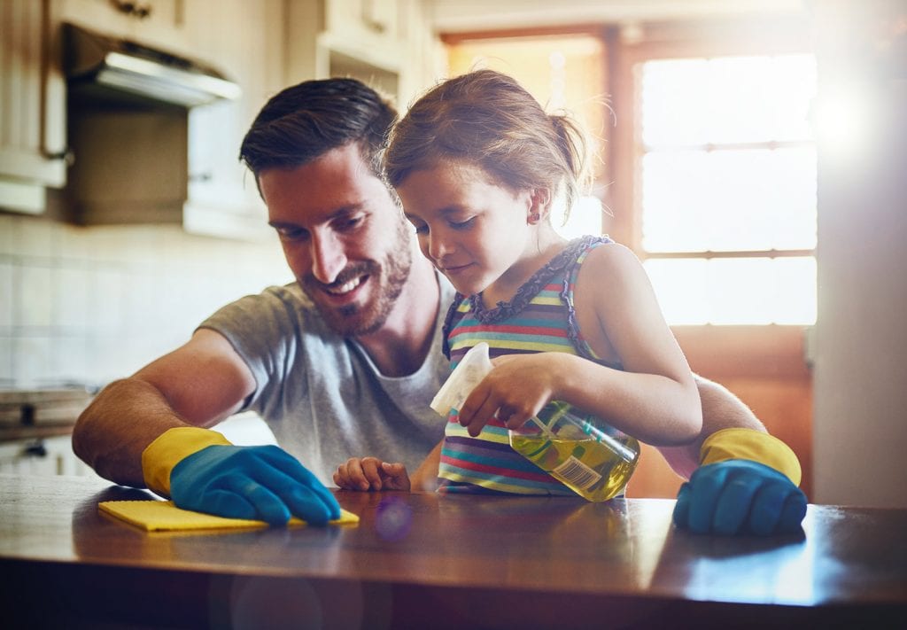 A father wearing blue and yellow cleaning gloves shows his yuong daughter how to clean the table. The little girl is holding a bottle of cleaning solution. 