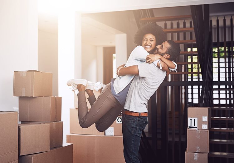 A young couple celebrates moving into a new home. The young man lifts his girlfriend up. The young woman smiles into the camera. 